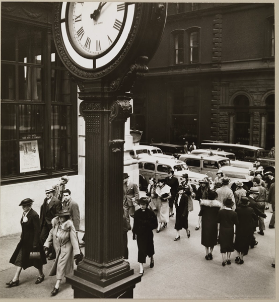 A group of people walk by a tall clock near a busy city street.