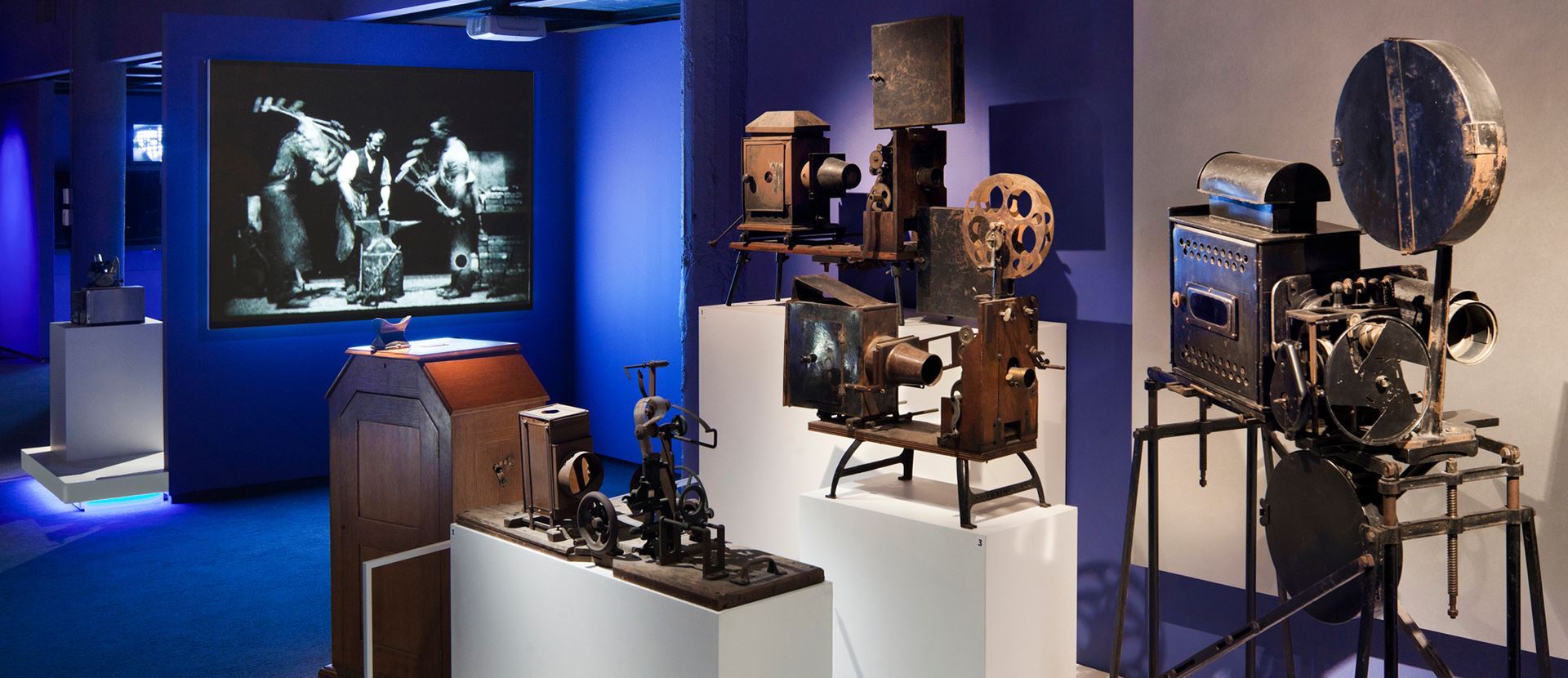 Image of 7 projectors in the Behind the Screen tour at the Museum of the Moving Image. The older projectors are rusty and on white platforms. A more modern projector hung from the ceiling projects a black and white film onto the wall of the dimly lit, blue-walled room.